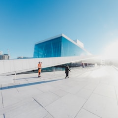 A modern building with a glass facade and an inclined roof. The sky is clear and blue. In the foreground, a person in orange safety clothing stands on the left side, while another person in black is walking towards the building.