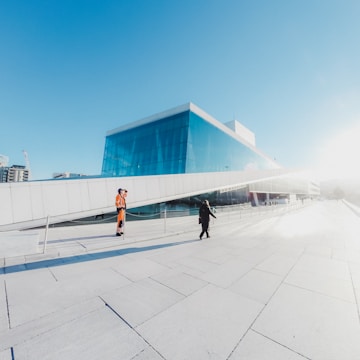 A modern building with a glass facade and an inclined roof. The sky is clear and blue. In the foreground, a person in orange safety clothing stands on the left side, while another person in black is walking towards the building.