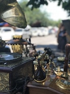 An antique setting displaying various vintage items including an old gramophone, a record player with a decorative brass horn, and numerous ornate brass and wooden objects. The scene appears to be outdoors, possibly at a market or fair, with several parked cars and trees visible in the background.