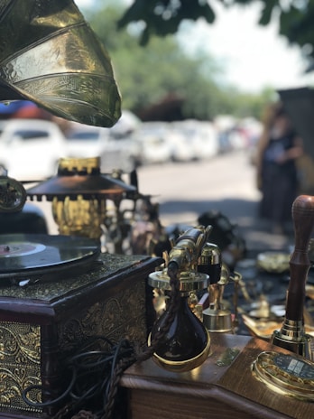 An antique setting displaying various vintage items including an old gramophone, a record player with a decorative brass horn, and numerous ornate brass and wooden objects. The scene appears to be outdoors, possibly at a market or fair, with several parked cars and trees visible in the background.