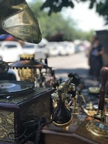 An antique setting displaying various vintage items including an old gramophone, a record player with a decorative brass horn, and numerous ornate brass and wooden objects. The scene appears to be outdoors, possibly at a market or fair, with several parked cars and trees visible in the background.