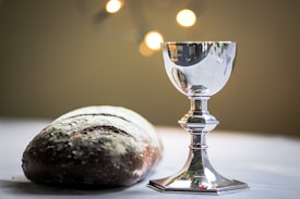 A silver chalice stands next to a rustic loaf of bread on a white cloth. Soft, glowing lights in the background create a warm ambiance.