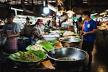 A bustling food market features vendors selling a variety of dishes. Large metal bowls and plates lined with banana leaves display an assortment of food items. Several people, including shoppers and vendors, interact amid the vibrant atmosphere. Overhead lamps provide illumination in the busy setting.