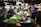 A bustling food market features vendors selling a variety of dishes. Large metal bowls and plates lined with banana leaves display an assortment of food items. Several people, including shoppers and vendors, interact amid the vibrant atmosphere. Overhead lamps provide illumination in the busy setting.