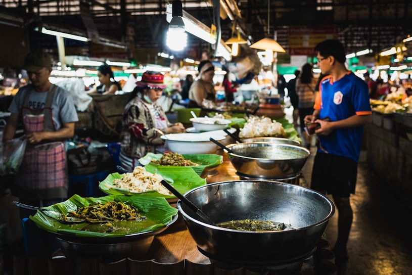 A bustling food market features vendors selling a variety of dishes. Large metal bowls and plates lined with banana leaves display an assortment of food items. Several people, including shoppers and vendors, interact amid the vibrant atmosphere. Overhead lamps provide illumination in the busy setting.
