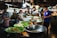 A bustling food market features vendors selling a variety of dishes. Large metal bowls and plates lined with banana leaves display an assortment of food items. Several people, including shoppers and vendors, interact amid the vibrant atmosphere. Overhead lamps provide illumination in the busy setting.