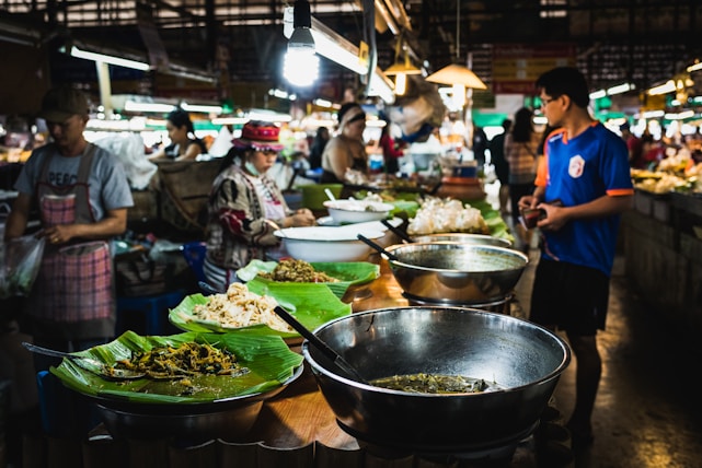 A bustling food market features vendors selling a variety of dishes. Large metal bowls and plates lined with banana leaves display an assortment of food items. Several people, including shoppers and vendors, interact amid the vibrant atmosphere. Overhead lamps provide illumination in the busy setting.