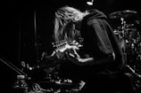 A black and white image of a musician kneeling on stage, playing an electric guitar. The person has long hair covering their face and is surrounded by musical equipment and drums in the background. A spotlight casts a dramatic shadow, highlighting the focused expression.