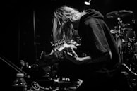 A black and white image of a musician kneeling on stage, playing an electric guitar. The person has long hair covering their face and is surrounded by musical equipment and drums in the background. A spotlight casts a dramatic shadow, highlighting the focused expression.