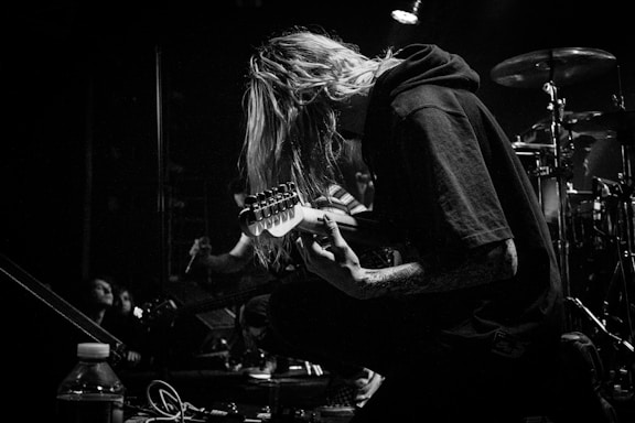 A black and white image of a musician kneeling on stage, playing an electric guitar. The person has long hair covering their face and is surrounded by musical equipment and drums in the background. A spotlight casts a dramatic shadow, highlighting the focused expression.