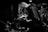 A black and white image of a musician kneeling on stage, playing an electric guitar. The person has long hair covering their face and is surrounded by musical equipment and drums in the background. A spotlight casts a dramatic shadow, highlighting the focused expression.