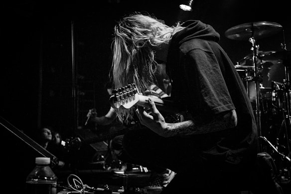A black and white image of a musician kneeling on stage, playing an electric guitar. The person has long hair covering their face and is surrounded by musical equipment and drums in the background. A spotlight casts a dramatic shadow, highlighting the focused expression.