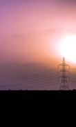 A silhouetted electricity pylon stands prominently against a vibrant sky during sunset or sunrise. The sky displays a gradient of warm colors ranging from purple to orange, creating a dramatic and serene atmosphere. Power lines extend from the pylon across the composition, adding structure and leading the viewer's eye into the distance.