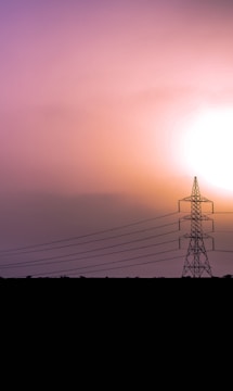 A silhouetted electricity pylon stands prominently against a vibrant sky during sunset or sunrise. The sky displays a gradient of warm colors ranging from purple to orange, creating a dramatic and serene atmosphere. Power lines extend from the pylon across the composition, adding structure and leading the viewer's eye into the distance.
