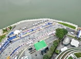 An aerial view of a large riverside event area featuring a racetrack layout with blue and yellow lines and numerous tents. People are scattered across the open space, some gathered in clusters. The river runs parallel to the event area, and there are green, park-like spaces with trees and architectural structures.