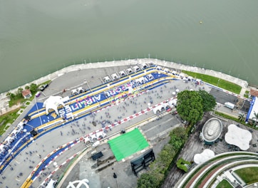 An aerial view of a large riverside event area featuring a racetrack layout with blue and yellow lines and numerous tents. People are scattered across the open space, some gathered in clusters. The river runs parallel to the event area, and there are green, park-like spaces with trees and architectural structures.