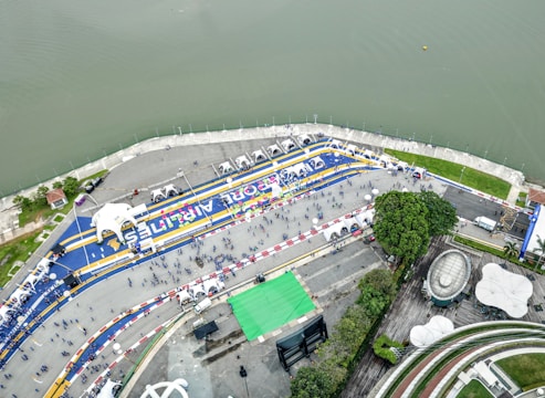 An aerial view of a large riverside event area featuring a racetrack layout with blue and yellow lines and numerous tents. People are scattered across the open space, some gathered in clusters. The river runs parallel to the event area, and there are green, park-like spaces with trees and architectural structures.