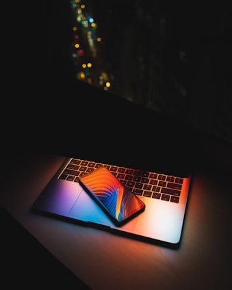 A laptop with illuminated keyboard creating colorful reflections. A smartphone with a vibrant screen displaying abstract graphics is placed on top of the laptop. The background is dark, with blurred city lights in the distance, emphasizing the contrast between technology and the night setting.