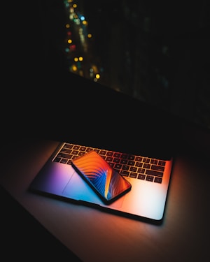 A laptop with illuminated keyboard creating colorful reflections. A smartphone with a vibrant screen displaying abstract graphics is placed on top of the laptop. The background is dark, with blurred city lights in the distance, emphasizing the contrast between technology and the night setting.