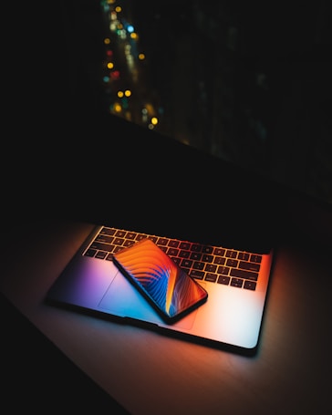 A laptop with illuminated keyboard creating colorful reflections. A smartphone with a vibrant screen displaying abstract graphics is placed on top of the laptop. The background is dark, with blurred city lights in the distance, emphasizing the contrast between technology and the night setting.
