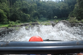 An off-road vehicle drives through a rocky stream, causing water to splash up dramatically. The vehicle's front bar and a red object are visible in the foreground, with lush green forest surrounding the scene.