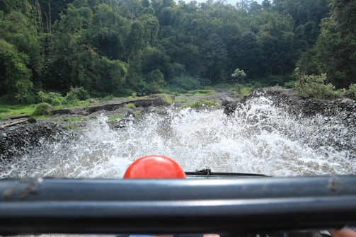 An off-road vehicle drives through a rocky stream, causing water to splash up dramatically. The vehicle's front bar and a red object are visible in the foreground, with lush green forest surrounding the scene.