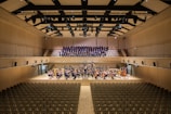 A spacious concert hall with rows of empty seats facing a stage. On the stage, a large orchestra is seated with various musical instruments, including strings and brass. Behind them, a choir stands in an elevated section, ready to perform. The hall has a modern architectural design with wood paneling and a high ceiling featuring lights and acoustic panels.