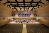 A spacious concert hall with rows of empty seats facing a stage. On the stage, a large orchestra is seated with various musical instruments, including strings and brass. Behind them, a choir stands in an elevated section, ready to perform. The hall has a modern architectural design with wood paneling and a high ceiling featuring lights and acoustic panels.