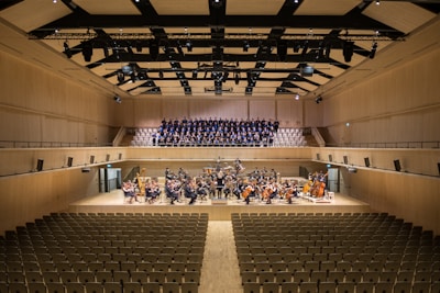 A spacious concert hall with rows of empty seats facing a stage. On the stage, a large orchestra is seated with various musical instruments, including strings and brass. Behind them, a choir stands in an elevated section, ready to perform. The hall has a modern architectural design with wood paneling and a high ceiling featuring lights and acoustic panels.