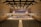 A spacious concert hall with rows of empty seats facing a stage. On the stage, a large orchestra is seated with various musical instruments, including strings and brass. Behind them, a choir stands in an elevated section, ready to perform. The hall has a modern architectural design with wood paneling and a high ceiling featuring lights and acoustic panels.