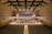 A spacious concert hall with rows of empty seats facing a stage. On the stage, a large orchestra is seated with various musical instruments, including strings and brass. Behind them, a choir stands in an elevated section, ready to perform. The hall has a modern architectural design with wood paneling and a high ceiling featuring lights and acoustic panels.