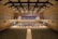 A spacious concert hall with rows of empty seats facing a stage. On the stage, a large orchestra is seated with various musical instruments, including strings and brass. Behind them, a choir stands in an elevated section, ready to perform. The hall has a modern architectural design with wood paneling and a high ceiling featuring lights and acoustic panels.