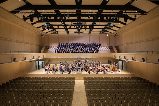 A spacious concert hall with rows of empty seats facing a stage. On the stage, a large orchestra is seated with various musical instruments, including strings and brass. Behind them, a choir stands in an elevated section, ready to perform. The hall has a modern architectural design with wood paneling and a high ceiling featuring lights and acoustic panels.