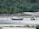 Two fishing boats are beached on a sandy shore, with lush green foliage and rocky outcrops in the background. The boats are tied with ropes to posts embedded in the sand, and the area appears to be at low tide with shallow water visible.