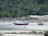 Two fishing boats are beached on a sandy shore, with lush green foliage and rocky outcrops in the background. The boats are tied with ropes to posts embedded in the sand, and the area appears to be at low tide with shallow water visible.