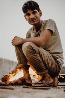 A man is crouched down, operating a handheld grinding tool that emits bright, flying sparks. The setting appears to be outdoors, and the man is wearing casual clothing, including a grey t-shirt and rolled-up trousers. His sandals are on the ground, and he is focused on the task at hand.