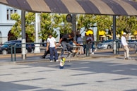 A group of people are in an outdoor urban setting, with one individual skillfully performing a skateboarding trick while others watch. The scene is lively, with sunlight casting shadows and trees in the background providing a scenic view. Cars are parked along the street, and people walk by, indicating a busy environment.