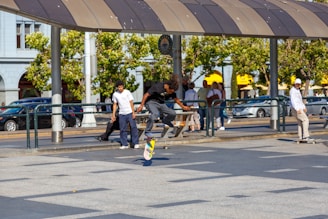 A group of people are in an outdoor urban setting, with one individual skillfully performing a skateboarding trick while others watch. The scene is lively, with sunlight casting shadows and trees in the background providing a scenic view. Cars are parked along the street, and people walk by, indicating a busy environment.