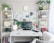 A well-organized home office setup featuring a white desk with a computer monitor displaying a succulent plant wallpaper. Shelves on both sides contain books, plants, headphones, decorative items, and storage boxes. A small white mirror sits on the desk alongside a few office supplies. A clock is mounted on the wall above the desk, and natural light filters in through sheer curtains, creating a bright and inviting atmosphere.