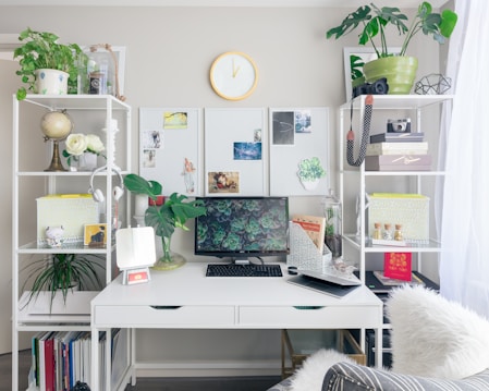 A well-organized home office setup featuring a white desk with a computer monitor displaying a succulent plant wallpaper. Shelves on both sides contain books, plants, headphones, decorative items, and storage boxes. A small white mirror sits on the desk alongside a few office supplies. A clock is mounted on the wall above the desk, and natural light filters in through sheer curtains, creating a bright and inviting atmosphere.