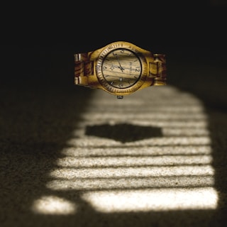 A wooden wristwatch appears to be floating above a surface with a pattern of light and shadow reminiscent of window blinds. The watch face displays black numerals and hands, with a detailed wood grain texture visible across both the face and band.
