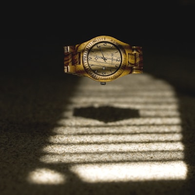 A wooden wristwatch appears to be floating above a surface with a pattern of light and shadow reminiscent of window blinds. The watch face displays black numerals and hands, with a detailed wood grain texture visible across both the face and band.