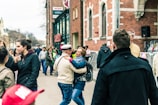 A lively street scene with people walking and interacting. A couple stands in the foreground, embracing and smiling. The background shows a busy street with groups of people conversing and walking past a brick building with large windows. Various individuals are dressed in casual clothing, and the scene conveys a sense of community and social interaction.