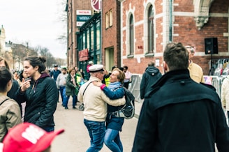 A lively street scene with people walking and interacting. A couple stands in the foreground, embracing and smiling. The background shows a busy street with groups of people conversing and walking past a brick building with large windows. Various individuals are dressed in casual clothing, and the scene conveys a sense of community and social interaction.