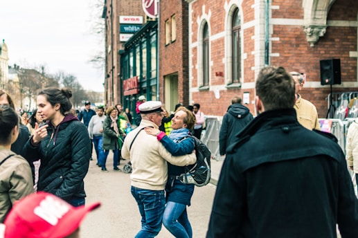 A lively street scene with people walking and interacting. A couple stands in the foreground, embracing and smiling. The background shows a busy street with groups of people conversing and walking past a brick building with large windows. Various individuals are dressed in casual clothing, and the scene conveys a sense of community and social interaction.