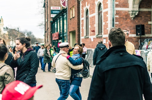 A lively street scene with people walking and interacting. A couple stands in the foreground, embracing and smiling. The background shows a busy street with groups of people conversing and walking past a brick building with large windows. Various individuals are dressed in casual clothing, and the scene conveys a sense of community and social interaction.