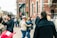 A lively street scene with people walking and interacting. A couple stands in the foreground, embracing and smiling. The background shows a busy street with groups of people conversing and walking past a brick building with large windows. Various individuals are dressed in casual clothing, and the scene conveys a sense of community and social interaction.