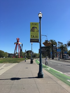 A sunny street scene features a cyclist riding on a designated green bike lane alongside a roadway. A tall street lamp displays a sign for the Port of San Francisco with an advertisement for water taxi landing. In the background, a large red sculpture and palm trees line the street, while several people are visible in the distance. The sky is clear and blue, enhancing the open and spacious atmosphere of the area.