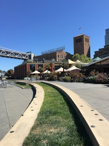A scenic urban setting featuring a modern bridge and brick industrial-style buildings. There is a green grass lawn with curving walkways lined by concrete borders. Several outdoor seating areas with tables and umbrellas are visible, along with greenery and small trees.