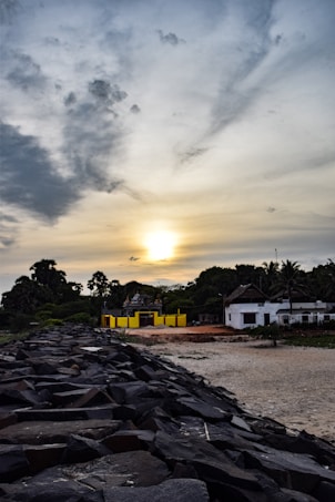 A dramatic sunset with a mix of dark clouds and clear sky creates a striking contrast. In the foreground, rocky embankments lead to a sandy shore. A small, colorful building with a yellow facade is nestled among green foliage. Palm trees and other greenery surround the area, and a few simple structures with thatched roofs are visible. The overall scene combines natural and man-made elements harmoniously.